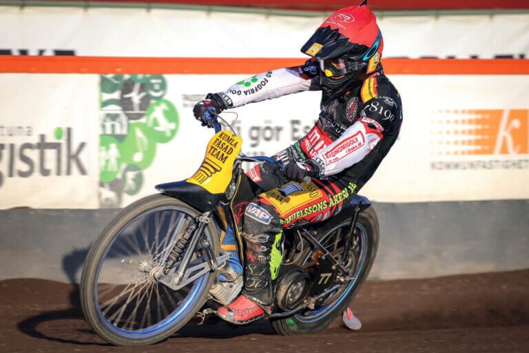 En med speedwayförare i full racingutrustning och hjälm lutar sig in i en kurva på en grusbana och sparkar upp damm. Motorcykeln och dräkten har olika sponsorlogotyper, och i bakgrunden syns hinder och speedwaybanereklam.
