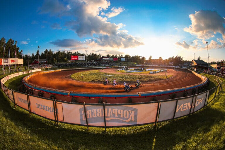 Fyra motorcyklister tävlar på en grusbana på en utomhusarena i solnedgången under evenemanget Rospiggarna 2025. Banan är omgiven av grönt gräs, banderoller och sittplatser, med moln och en blå himmel i bakgrunden.
