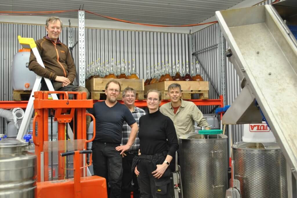 Fem personer står och ler inne i en metallväggig anläggning i Lohärad med bryggutrustning, stora stålbehållare, glasflaskor och industrimaskiner och tillverkar hantverksmässig cider av färsk äppelodling.