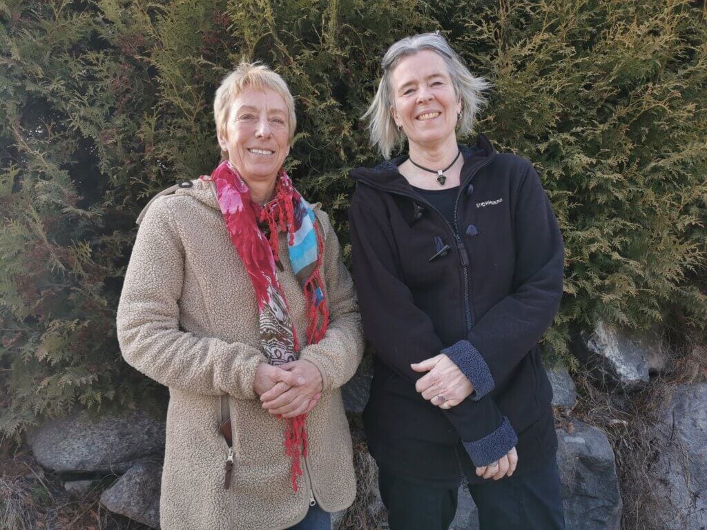 Två vän står utomhus framför gröna buskar, båda ler mot kameran. Den ena bär en brun kappa och en färgglad halsduk, den andra en svart jacka. Stenar syns längst ner i bilden och fångar ett ögonblick från livets sista kapitel.