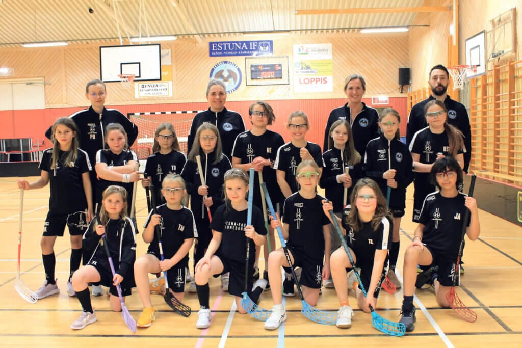 Ett ungdomslag i innebandy från Norrtälje poserar för ett gruppfoto i ett inomhusgym som utstrålar idrottsglädje. Flickorna i svarta uniformer håller stolt i sina innebandyklubbor och bakom dem står fyra engagerade själ-tränare bland nät och sportutrustning.