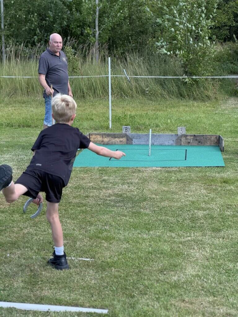 En ung pojke i svarta shorts och t-shirt kastar en hästsko mot ett mål, medan en äldre man tittar på på en gräsplan. Det finns siffror och en grön matta vid målet, vilket skapar härlig gemenskap runt en pinne.