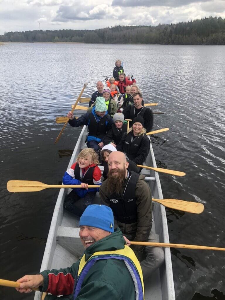 En grupp människor, både vuxna och barn, sitter tätt tillsammans i en lång kanot på en lugn sjö och njuter av drakrodd. Var och en håller i en paddel och bär flytväst, med träd och en molnig himmel i bakgrunden.