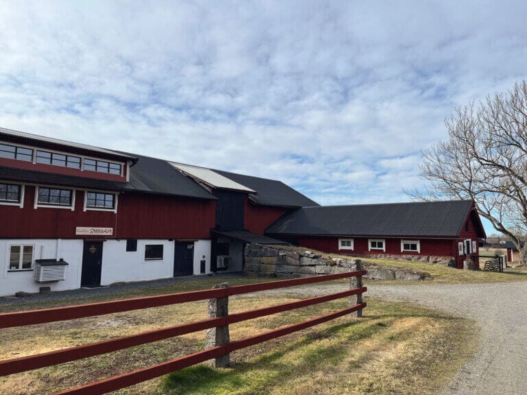En stor röd trälada, känd som Ingolfs ladugård, med vita snickerier står på en gräsbevuxen tomt som avgränsas av ett trästaket och en grusgång. Ett lövlöst träd och en delvis molnig himmel skapar en inspirerande vy för konstnärer att växa kreativt.