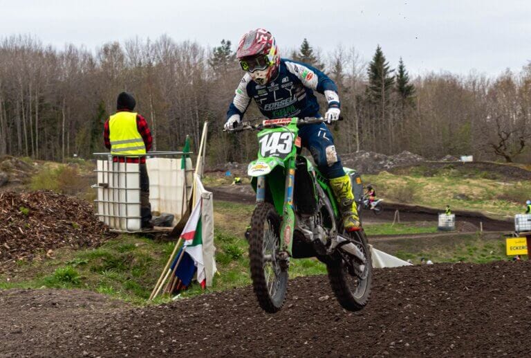 En motocrossförare iförd utrustning och hjälm, nummer 143, kör en grön MK Orion dirt bike över en kulle på en grusbana utomhus. En person i gul väst står i närheten. Träd och den mulna himlen 2025 finns i bakgrunden.