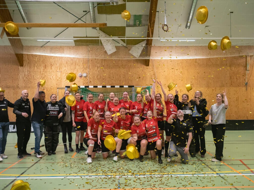 En grupp Midas damer i röda och svarta sportuniformer firar inomhus och kastar guldhattar i luften medan guldkonfetti faller. De står på en grön plan med nät och träväggar och firar sin seger i division 2.