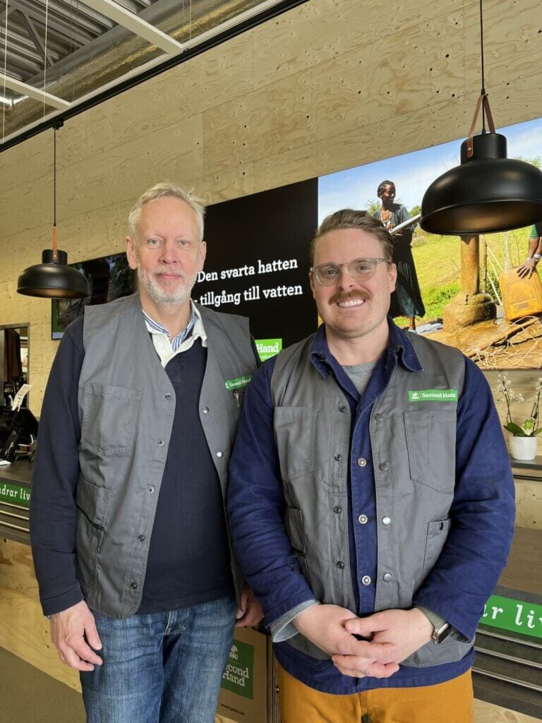 Två människor i grå västar står sida vid sida inomhus framför en träbänk och en väggdisplay med text, ett PMU-foto av en person utomhus och hängande lampor ovanför dem.