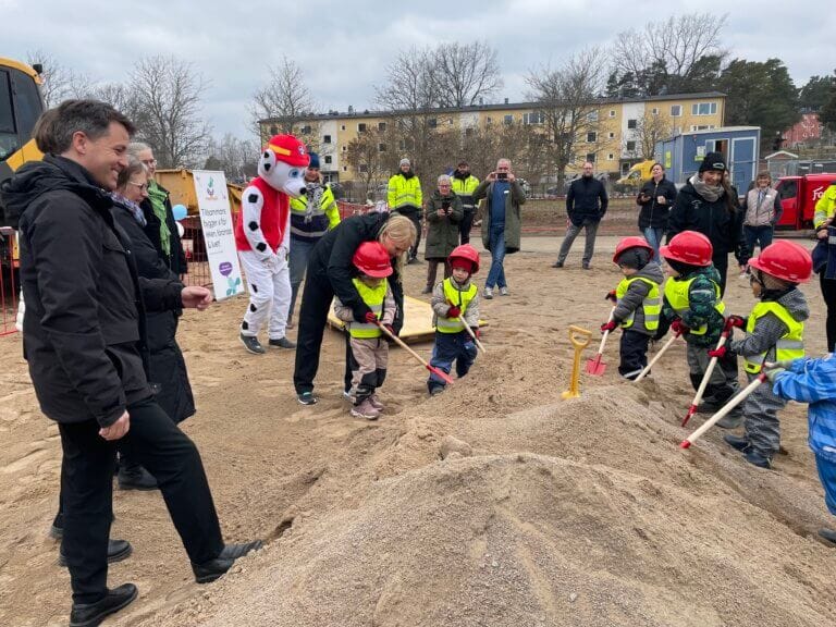 En grupp barn i skyddsvästar och röda hjälmar gräver i en sandhög på byggarbetsplatsen Rimbo ny förskola, omgivna av vuxna, fotografer och en person i hundmaskotkostym.