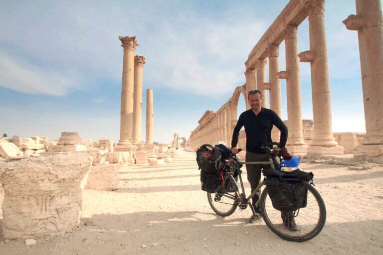 En man, som påminner om upptäcktsresenär Lars Bengtsson, står bredvid sin fullastade touringcykel bland gamla stenpelare och ruiner i en ökenmiljö, redo att cykla 120000 kilometer under en klar himmel.