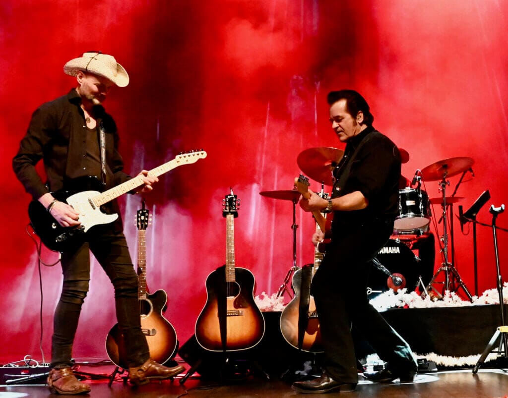 Två musiker från Opalen uppträder i Rock n' Country Parade och spelar elgitarr på en scen med röd belysning och rök. Båda klädda i svart, den ene med cowboyhatt, står de framför ett trumset och tre akustiska gitarrer.