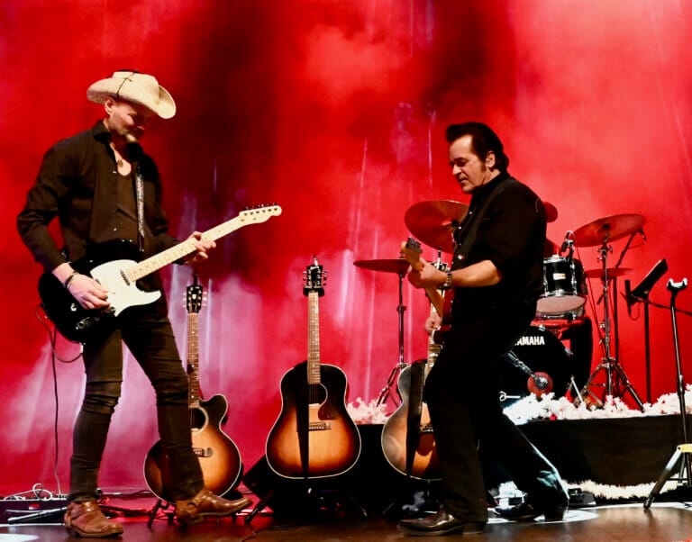 Två musiker från Opalen uppträder i Rock n' Country Parade och spelar elgitarr på en scen med röd belysning och rök. Båda klädda i svart, den ene med cowboyhatt, står de framför ett trumset och tre akustiska gitarrer.