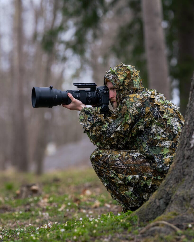 Djurfotografen Patrik Luther hukar bakom ett träd i kamouflagekostym och siktar på den perfekta bilden med sin proffskamera och stora teleobjektiv i en skogig utomhusmiljö.