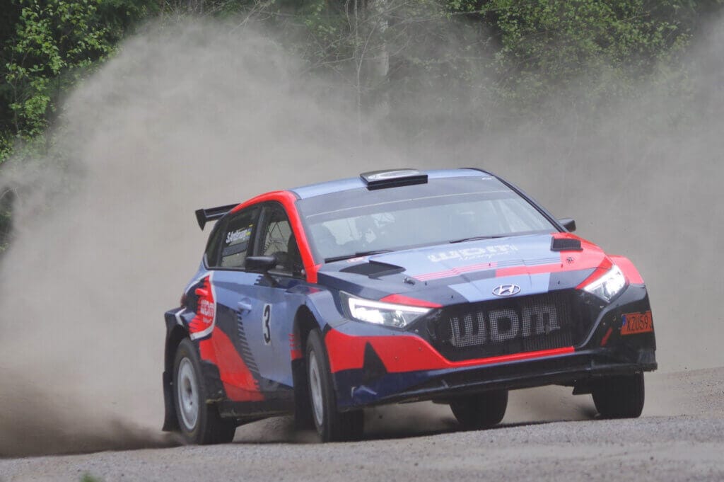 En blå och röd Andervang-rallybil med nummer 3 på sidan driver runt ett grusvägshörn och sparkar upp damm under ett spännande minnesrally, med gröna träd i bakgrunden.