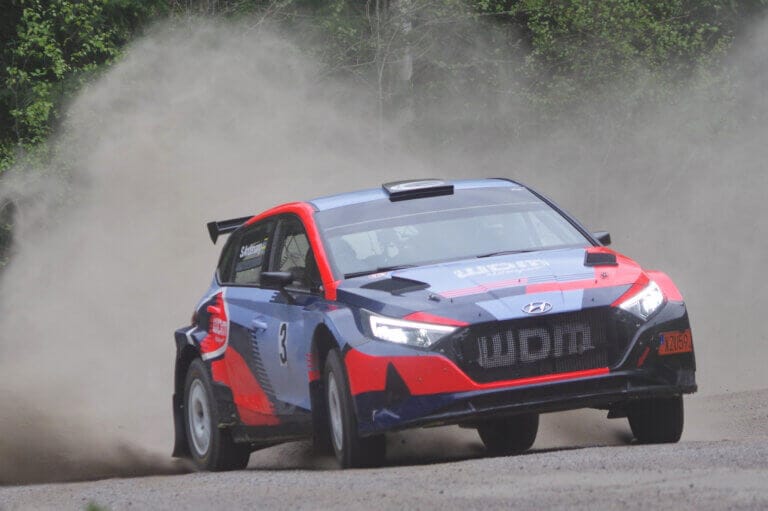 En blå och röd Andervang-rallybil med nummer 3 på sidan driver runt ett grusvägshörn och sparkar upp damm under ett spännande minnesrally, med gröna träd i bakgrunden.