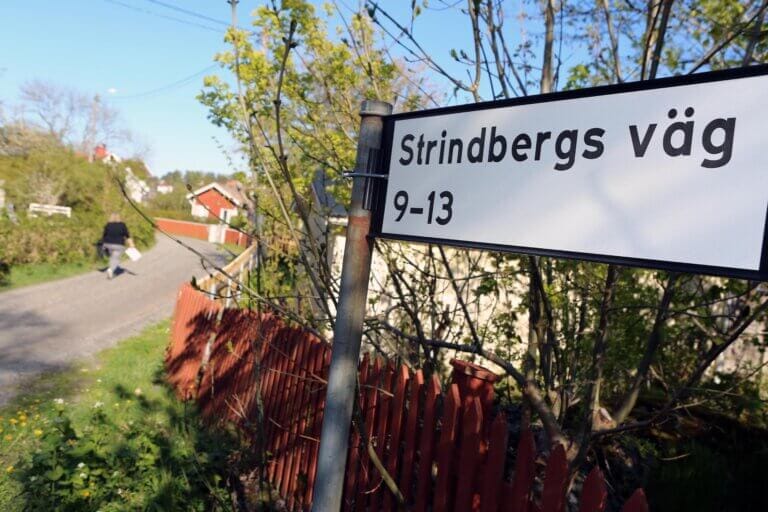 Närbild av en gatuskylt med texten "Strindbergs väg 9-13" intill ett rött staket i Furusund, med träd och en person som vandrar längs en grusgång mot röda hus i bakgrunden.