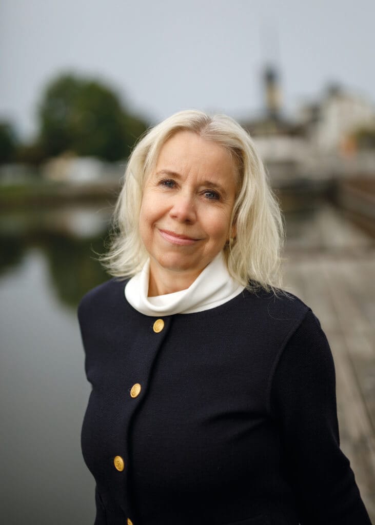 En kvinna med axellångt blont hår och ett lätt leende står vid ett vattendrag och funderar över skrivandet och överlevnadens höga pris. Hon bär en mörk kavaj med guldknappar, träd och byggnader skymtar mjukt i bakgrunden.