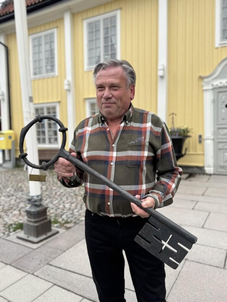 En man i rutig skjorta står utomhus på en kullerstensbelagd gång och håller i Norrtälje nyckel. Bakom honom står en gul träbyggnad med vita lister och en grå dörr som påminner om Knutte Alms tid.