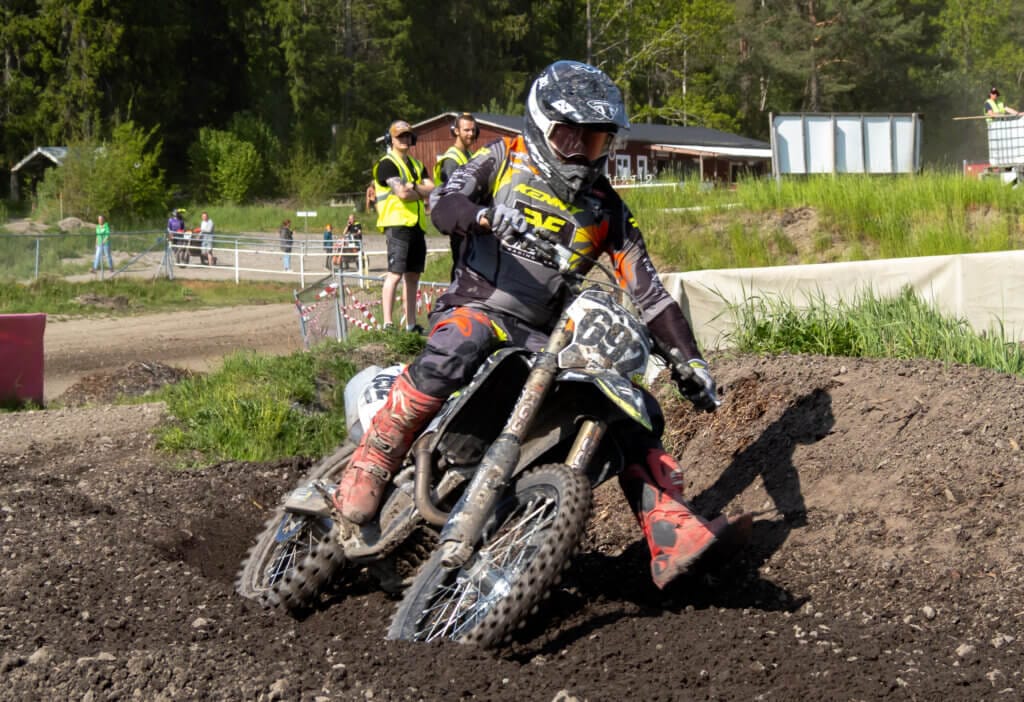 En motocrossförare med hjälm och skyddsutrustning kör runt ett hörn på en grusbana på en motorcykel under Orion Pokalen och sparkar upp jord. Flera personer står i bakgrunden nära grönt gräs och träd.