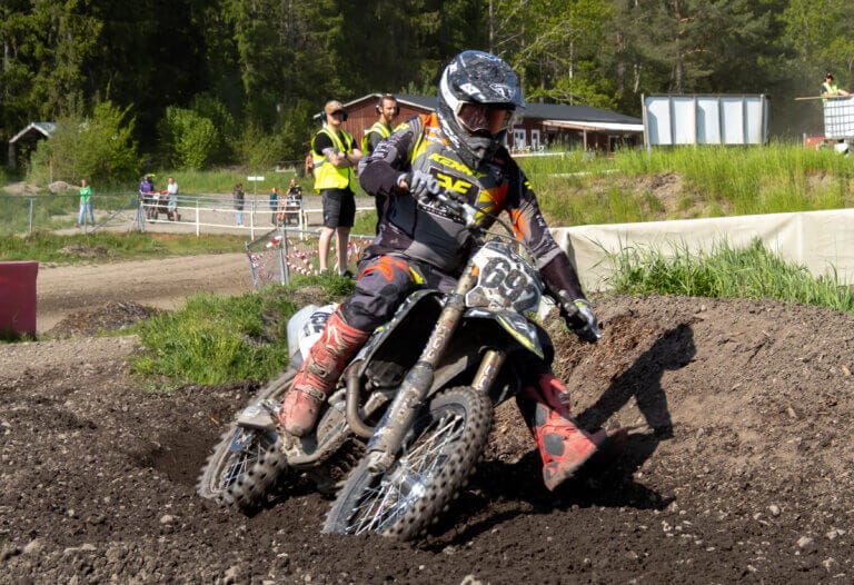En motocrossförare med hjälm och skyddsutrustning kör runt ett hörn på en grusbana på en motorcykel under Orion Pokalen och sparkar upp jord. Flera personer står i bakgrunden nära grönt gräs och träd.