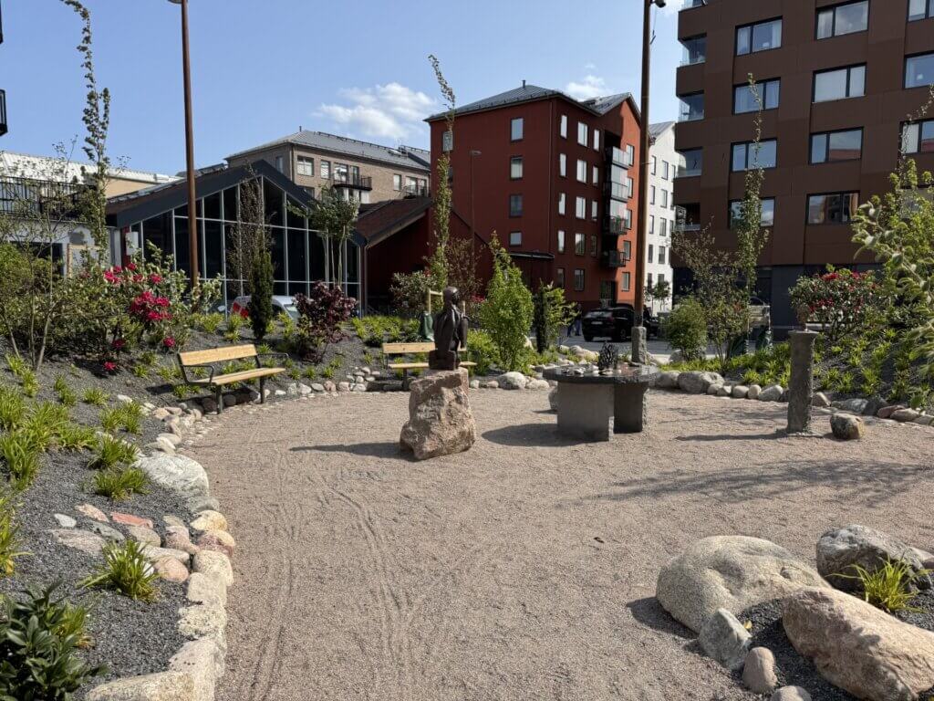 En liten urban ny park med grusmark, omgiven av stenar och grönska, har två träbänkar och en central staty på en sten, med moderna hyreshus i bakgrunden under en klar himmel.