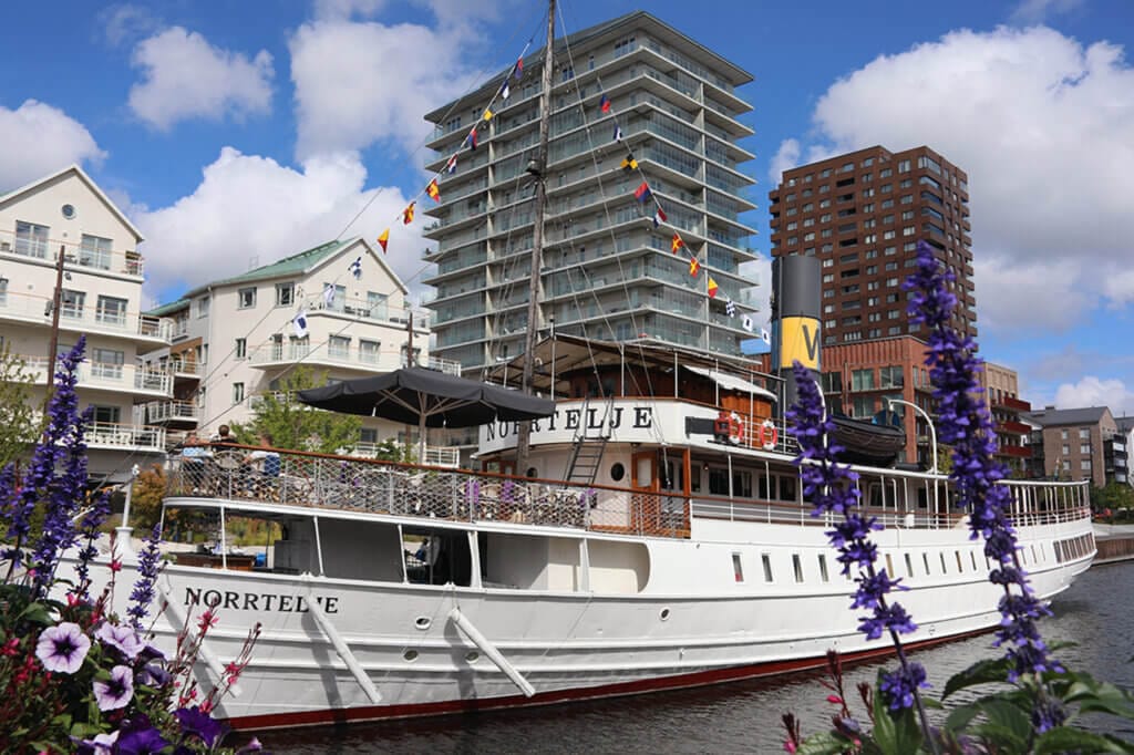En vit båt vid namn Norrtälje, firar 125 år som Skärgårdens drottning, ligger förtöjd vid en kanal med höga moderna lägenheter bakom. Lila blommor ramar in förgrunden under en ljus, delvis molnig dag.