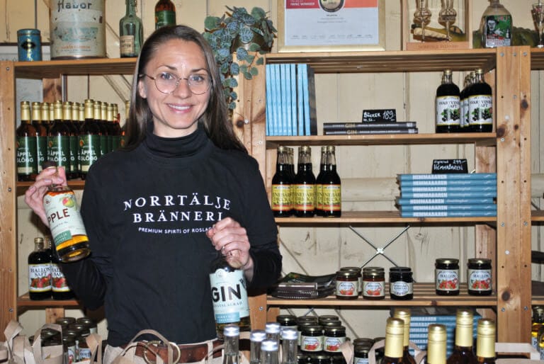 En kvinna med glasögon och en svart "Norrtälje Bränneri"-skjorta, dryckeshantverkare, håller i flaskor med äppelbrännvin och gin och står vid hyllor fyllda med alkoholdrycker, konserver och böcker i en livlig butik.