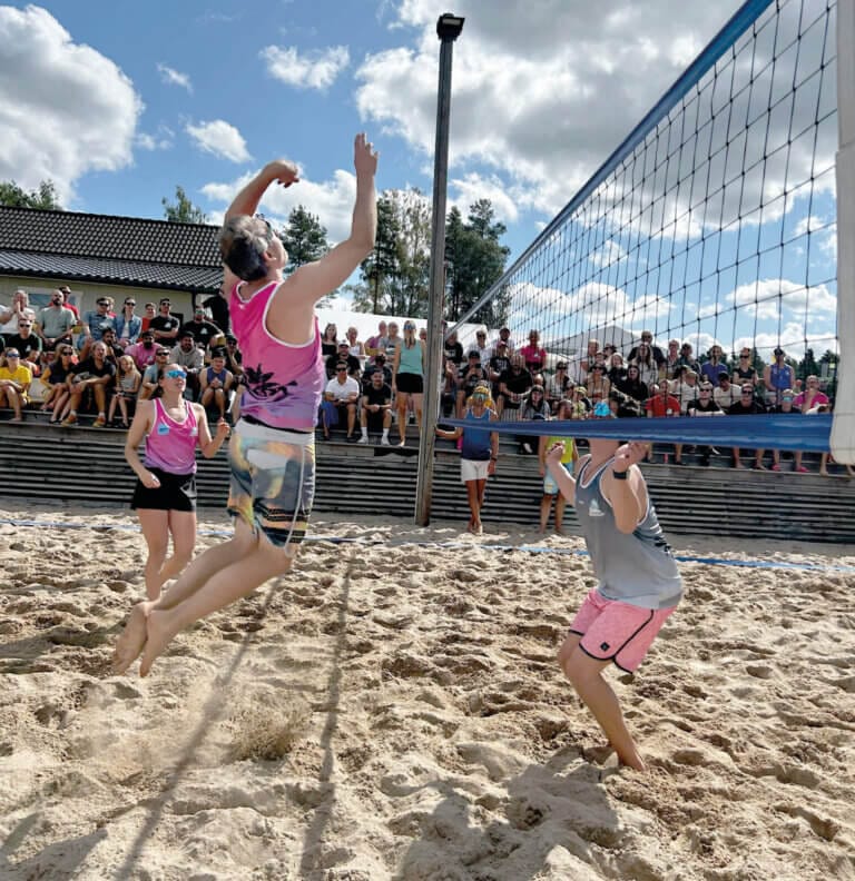 Två personer spelar beachvolleyboll på en sandplan under en turnering, med en publik som tittar på från läktare. En spelare hoppar för att slå bollen medan den andra förbereder sig för att blockera, allt en del av kampen mot cancer denna soliga dag med spridda moln.
