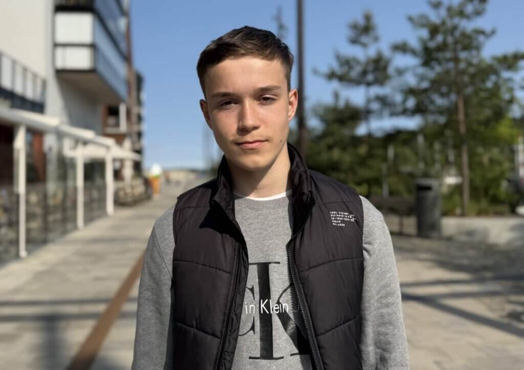 Eric drömmer om Hollywood när han står utomhus på en solbelyst trottoar, klädd i en svart väst över en grå Calvin Klein-sweatshirt. Träd och byggnader syns i bakgrunden under en klarblå himmel.