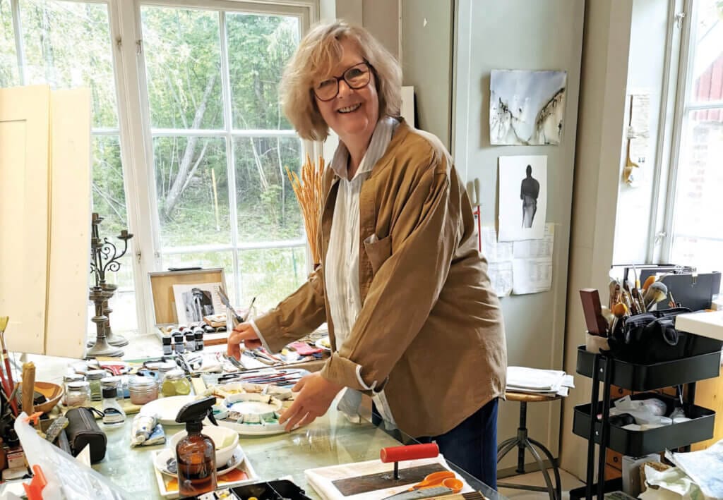 En kvinna med glasögon och ljust hår ler medan hon arbetar med ett konstprojekt vid ett bord i en ljus studio, omgiven av konstmaterial, medan grönskan utanför antyder att det är åter dags för den uppskattade konstrundan.