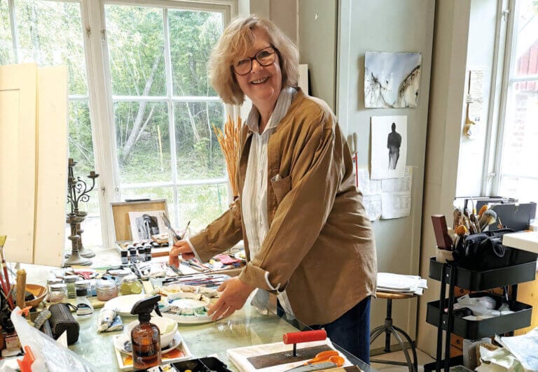 En kvinna med glasögon och ljust hår ler medan hon arbetar med ett konstprojekt vid ett bord i en ljus studio, omgiven av konstmaterial, medan grönskan utanför antyder att det är åter dags för den uppskattade konstrundan.