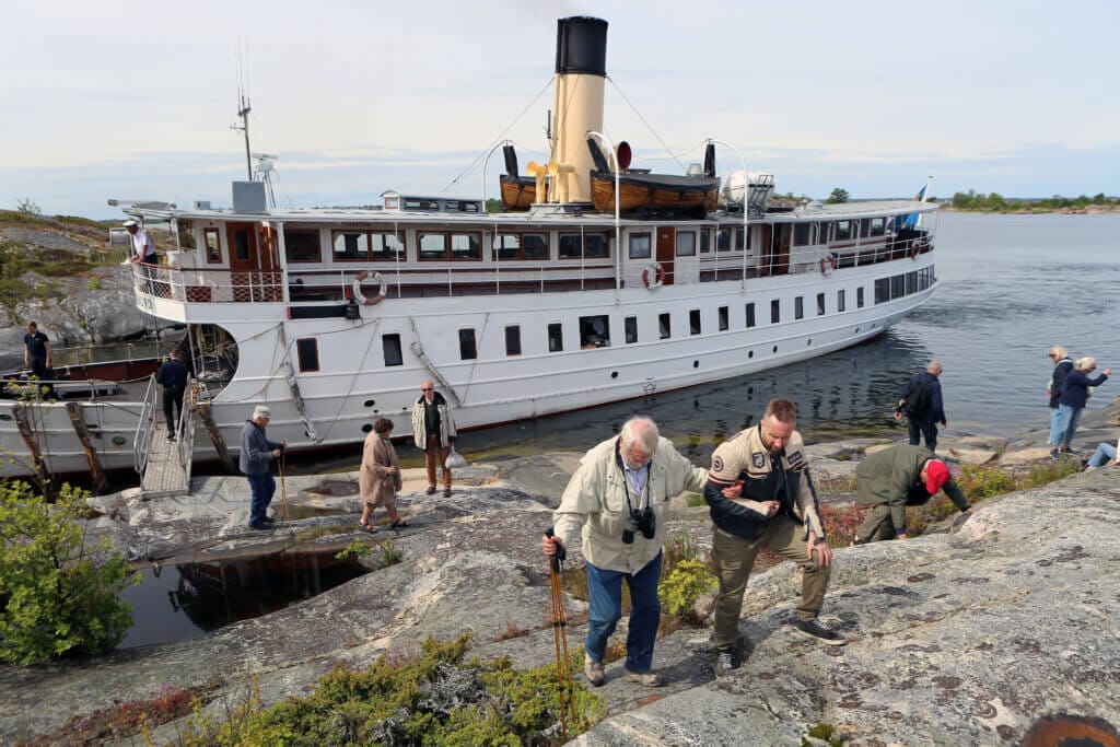 En grupp människor går i land från en stor vit båt som ligger vid Himmelskärs steniga, romantiska landskap. Flera går på klipporna och några fotograferar eller använder vandringsstavar.