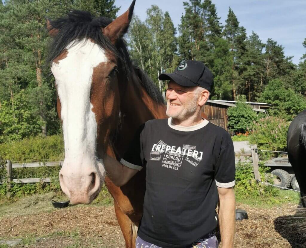 En man med svart keps, svart t-shirt och mönstrade shorts står utomhus vid en brun och vit häst -ingen hov syns. Träd och ett trästaket finns i bakgrunden en solig dag.