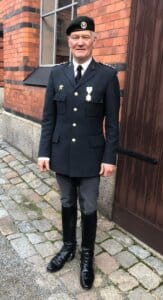 En man i en formell militäruniform med medaljer står på en kullerstensbelagd stig bredvid en tegelbyggnad med stora fönster och en hög trädörr-ingen hov, ingen häst, men han bär glänsande svarta stövlar, handskar och en svart basker.