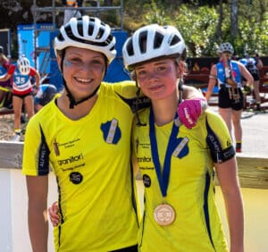 Två unga idrottare i gula Häverödals SK-tröjor och vita hjälmar står sida vid sida och ler mot kameran. Den ena bär stolt ett medaljregn från JSM. Andra tävlande och sportutrustning syns i bakgrunden.