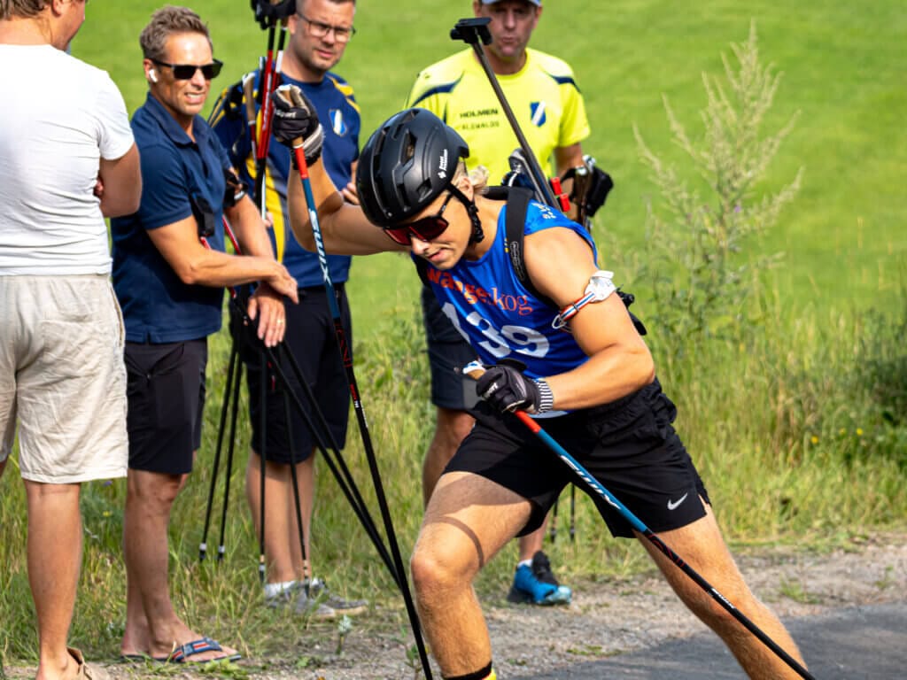 En person i sportkläder och hjälm tävlar i JSM rullskidor, representerande Häverödals SK, med hjälp av skidstavar på en asfalterad bana. Åskådare och funktionärer står i närheten med kameror eller solglasögon. Gräs och grönska syns i bakgrunden.