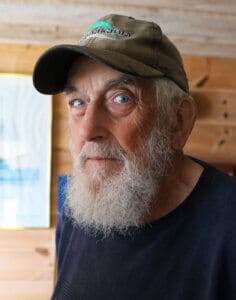 En äldre man med vitt skägg och keps står inomhus i ett trärum fyllt med olika föremål, bland annat ett automatiskt utkast, en inramad skeppsbild och ljus på hyllor i bakgrunden.