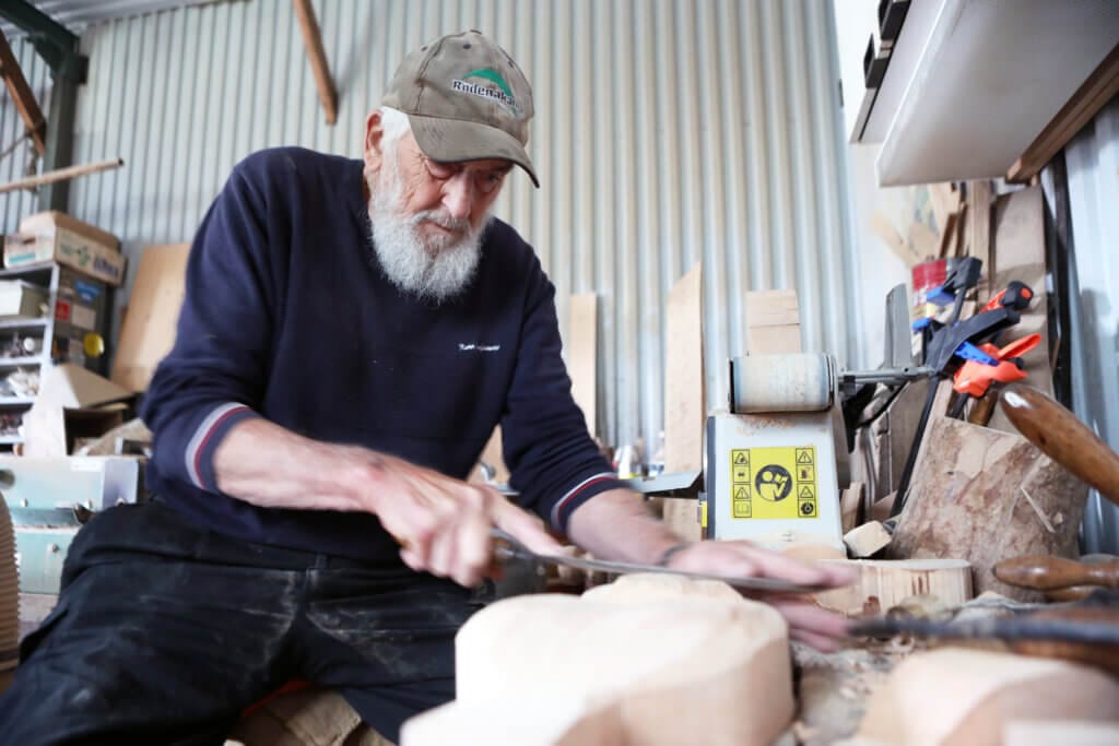En äldre man med vitt skägg och keps använder en rasp för att forma trä vid en rörig arbetsbänk i sin verkstad med väggar av korrugerad metall. Olika verktyg och träbitar ligger utspridda runt honom och skapar en autentisk, automatiskt utkastad atmosfär.