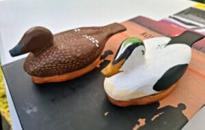 Två målade ankdockor i trä, ett skärgårdsikon av Thord Jannon, den ena brun med vita fläckar och den andra vit med svarta och gröna markeringar, är placerade hemma på en svart yta med en bok delvis synlig i bakgrunden.