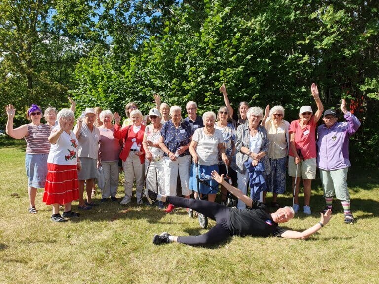 En grupp äldre på ett seniorkollo står utomhus framför gröna träd och ler och vinkar till kameran. En person ligger på gräset framför med armar och ben utsträckta och firar en solig jubileumsdag.