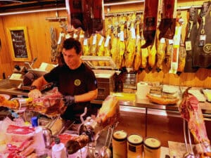En man bakom en disk skivar skinka i en butik, där vin och mattor visas bland delikatessutrustning och förpackade produkter, medan flera hela skinkor hänger på träväggen.