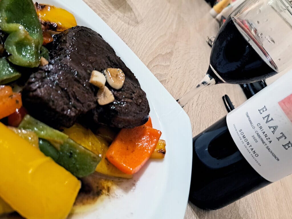 En tallrik med en bit biff toppad med vitlök, serverad med paprika och morötter, vilar på ett träbord bredvid ett glas och en flaska Enate vin.