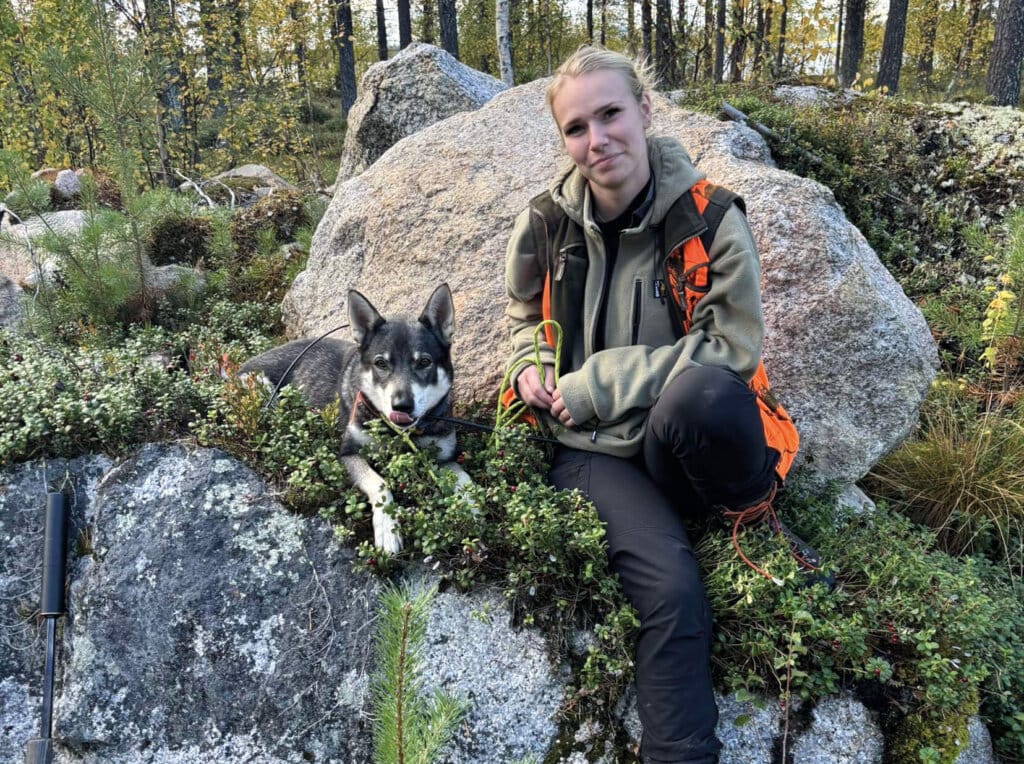 En jägare i friluftskläder sitter på en stor sten i ett skogsområde, bredvid en av hennes hundarna som ligger på den mossiga marken. Träd och grönska omger dem, och ett gevär syns delvis i förgrunden.