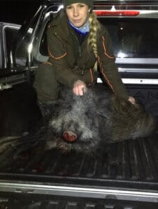 En jägare i friluftskläder står på knä bredvid ett vildsvin som ligger på flaket till en pickup på natten och håller djurets huvud efter en lyckad jakt.