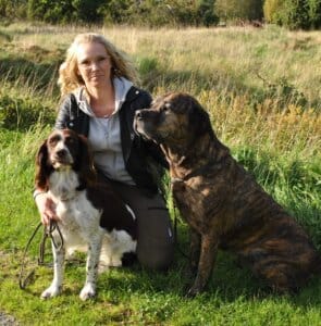 En person med glasögon står på knä i gräset utomhus och håller i kopplen till två hundarna: en vit och brun hund och en stor brindle hund. Grönt gräs och lövverk fyller bakgrunden.