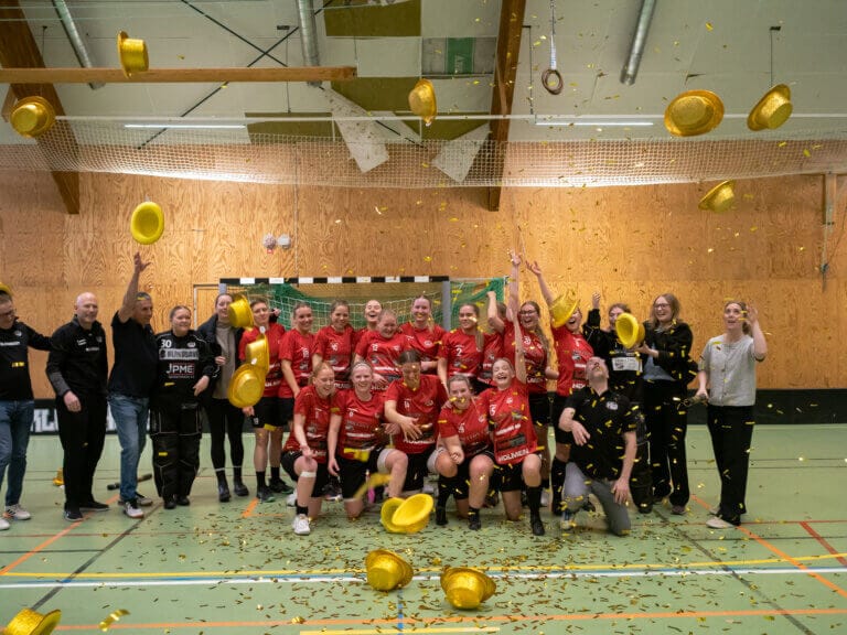 Ett damidrottslag i röda uniformer firar på en inomhusplan, omgivet av tränare och personal. När guldhattar och konfetti fyller luften jublar de glatt - som stjärnor i tv-serier som firar sitt stora premiärdatum.