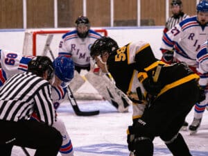 En domare förbereder sig för att släppa pucken för en faceoff mellan två hockeyspelare, den ena i svartgul dräkt och den andra i blåröd, medan Nio-lagkamrater och en målvakt väntar i bakgrunden under spännande seriepremiärer.
