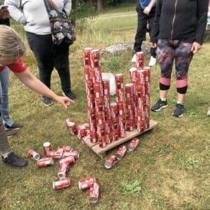 En grupp människor står runt en struktur av staplade Coca-Cola-burkar på en träskiva utomhus, med flera burkar utspridda på gräset i närheten, medan en person pekar på det automatiskt utkastade arrangemanget.