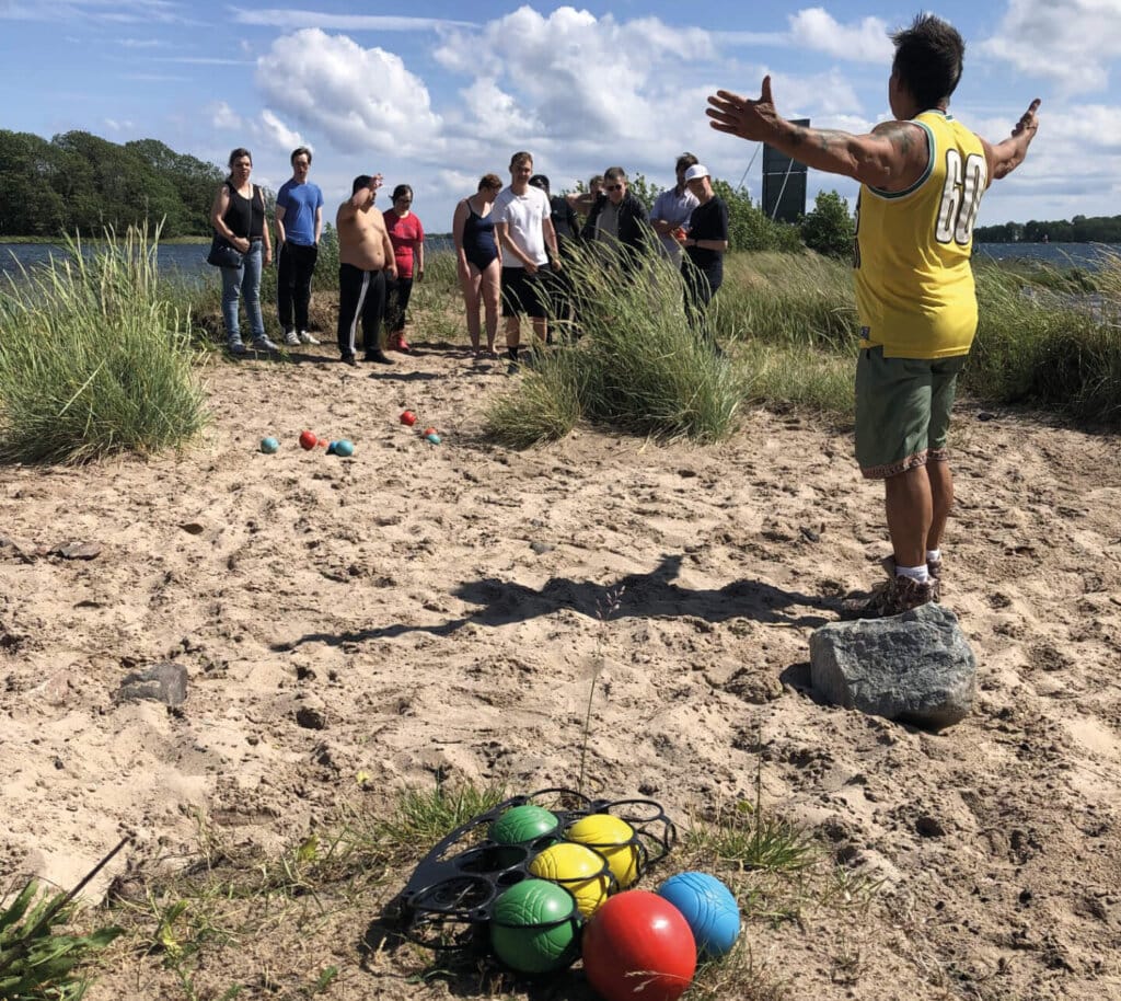 En man i gul tröja står med utsträckta armar på en sandyta vid vattnet och leder en grupp vänner i ett äventyr. Bocciabollar och annan sportutrustning ligger utspridda i närheten, och gräs och blå himmel kompletterar bilden.