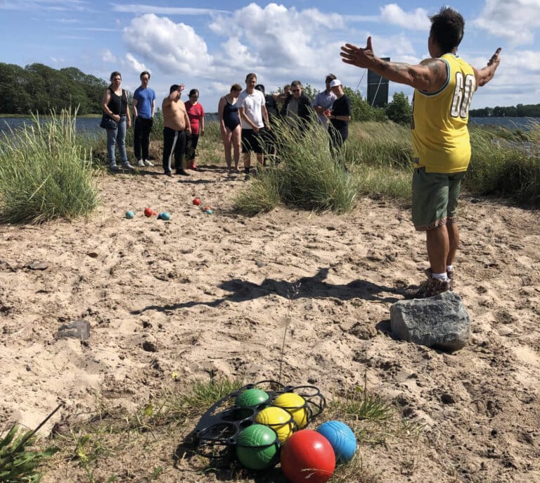 En man i gul tröja står med utsträckta armar på en sandyta vid vattnet och leder en grupp vänner i ett äventyr. Bocciabollar och annan sportutrustning ligger utspridda i närheten, och gräs och blå himmel kompletterar bilden.