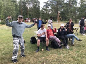 En grupp vänner, några sittande på en bänk och några stående, poserar lekfullt i ett gräsbevuxet utomhusområde med träd och röda trästugor i bakgrunden och fångar andan av äventyr.